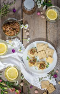 A rustic table setting with a plate of crackers, cheese, and a spread topped with herbs, accompanied by Ploughmans Pickle, mason jars of lemon water, a jar of chutney, flowers, and a white cloth napkin.
