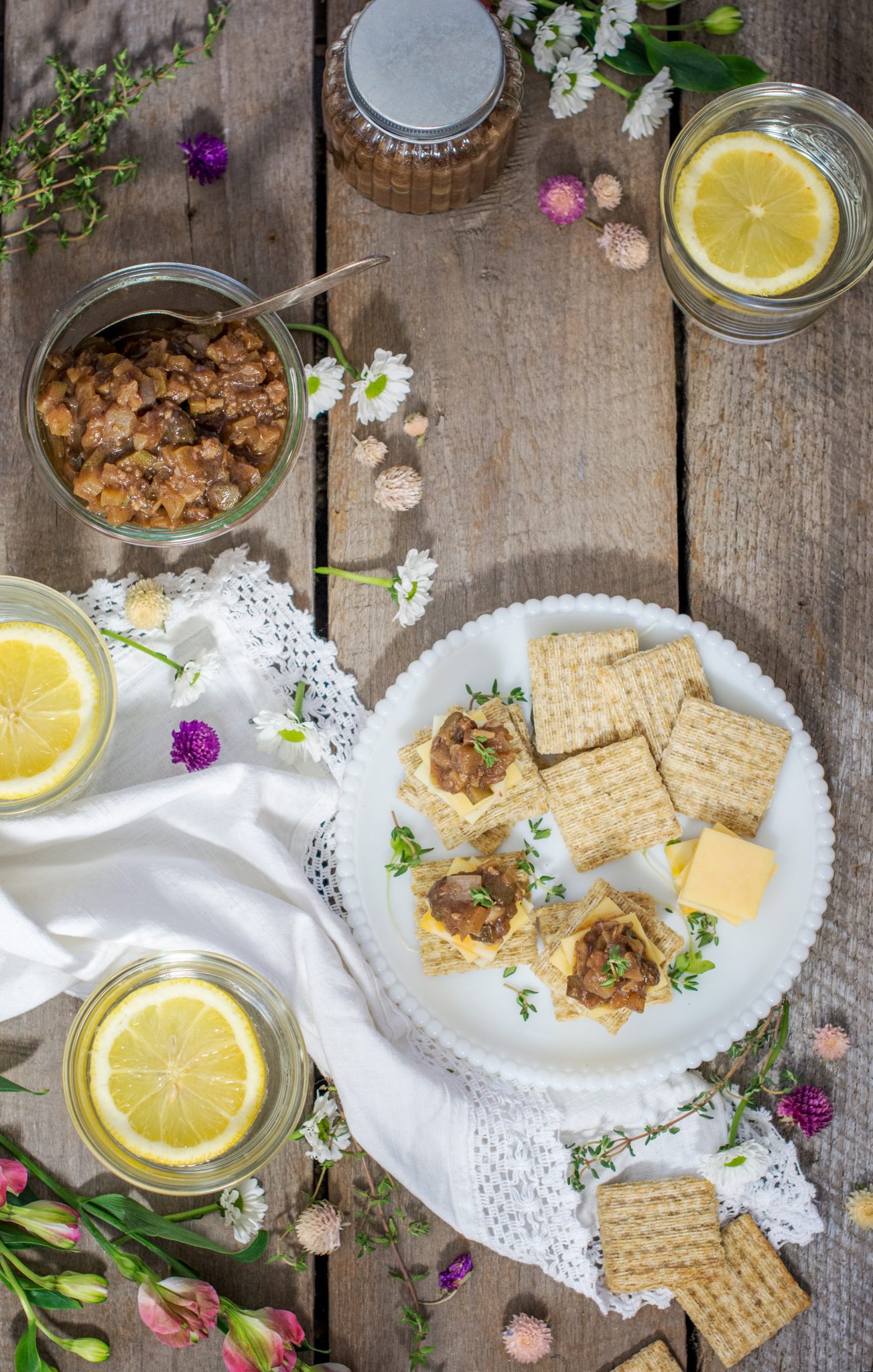 A rustic table setting with a plate of crackers, cheese, and a spread topped with herbs, accompanied by Ploughmans Pickle, mason jars of lemon water, a jar of chutney, flowers, and a white cloth napkin.