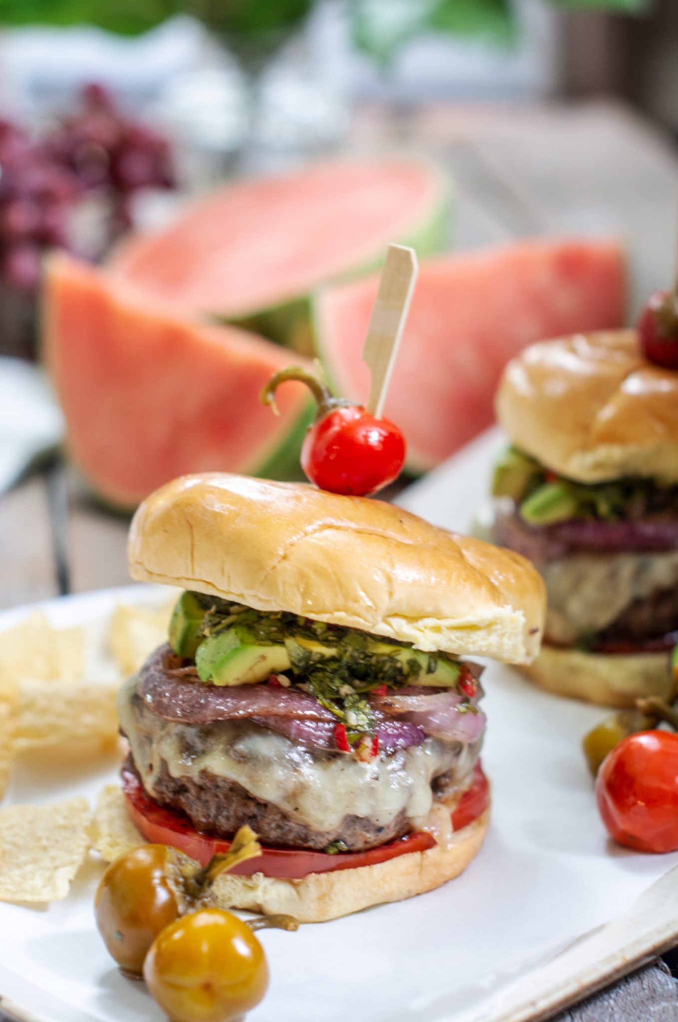 Two cheeseburgers topped with avocado, greens, and a cherry tomato skewer on brioche buns sit on a plate with tortilla chips. Inspired by our Chimichurri Burger Recipe, sliced watermelon and more tomatoes are in the background.