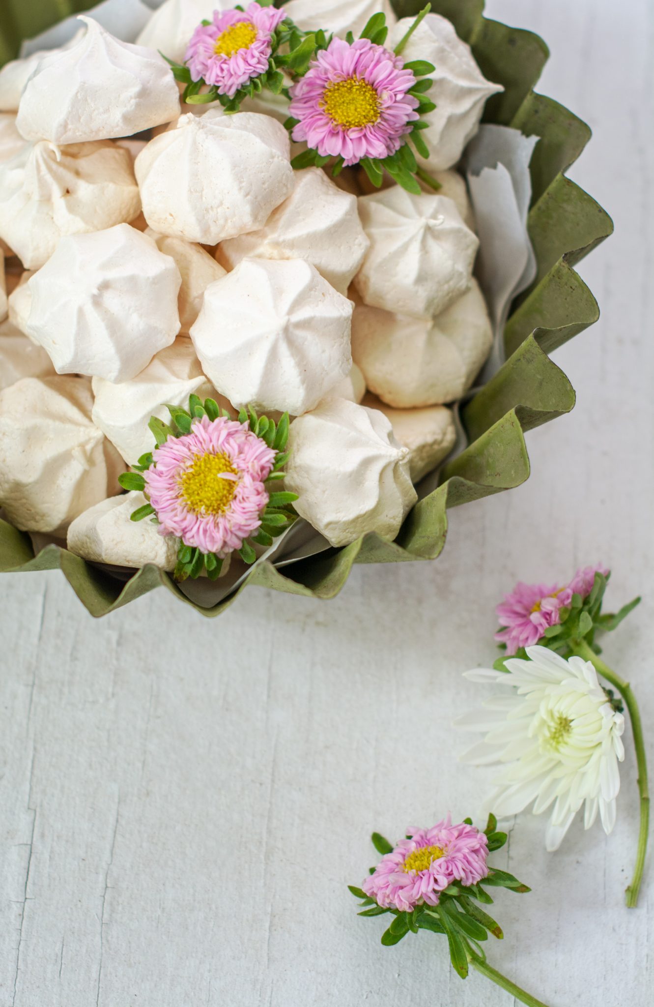 A green bowl filled with easy meringue cookies, decorated with pink and white daisy-like flowers on top and beside the bowl, set on a light wooden surface.