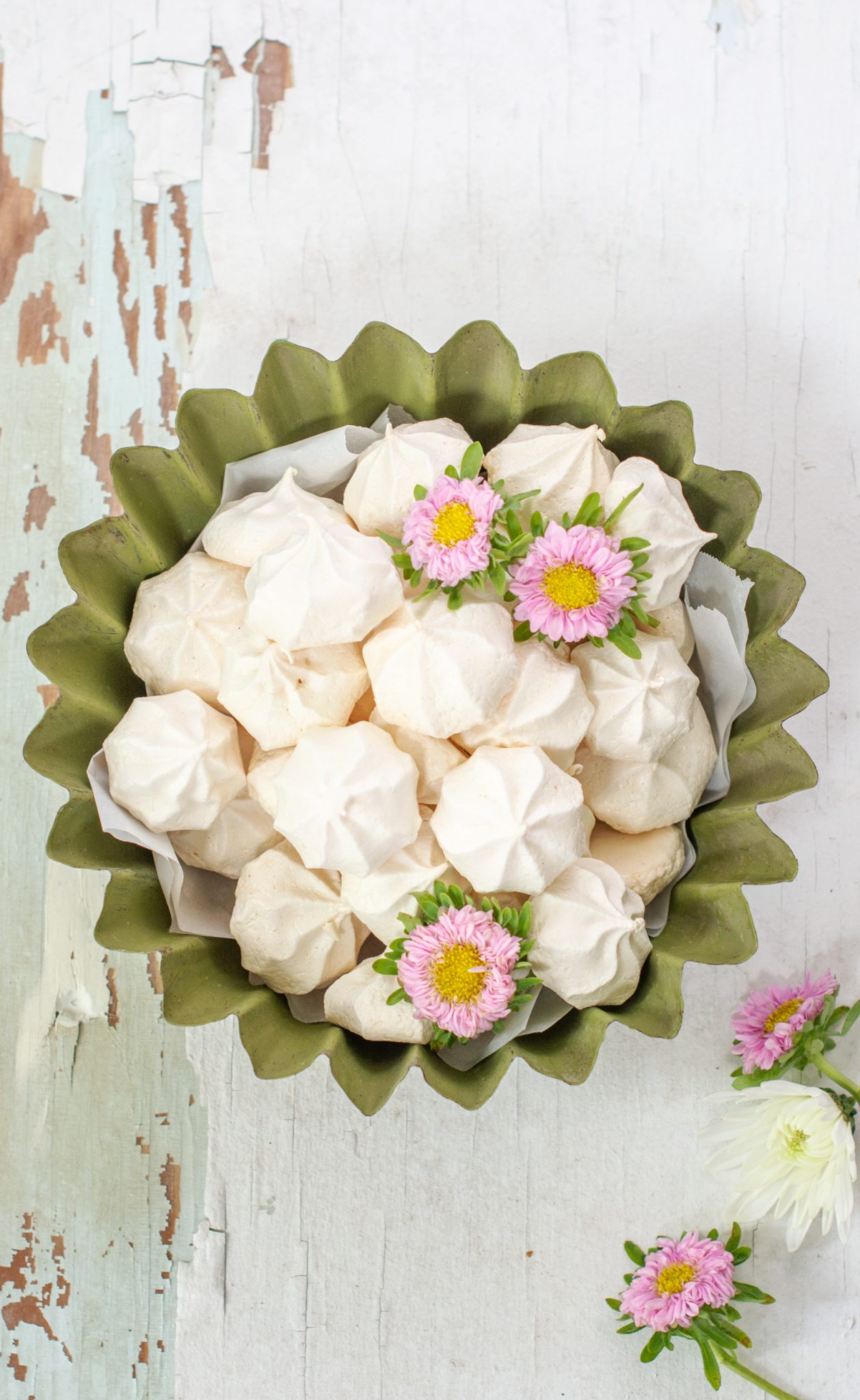 A green fluted bowl filled with easy meringue cookies, decorated with small pink and white daisy-like flowers, sits on a rustic white wooden surface with peeling paint.