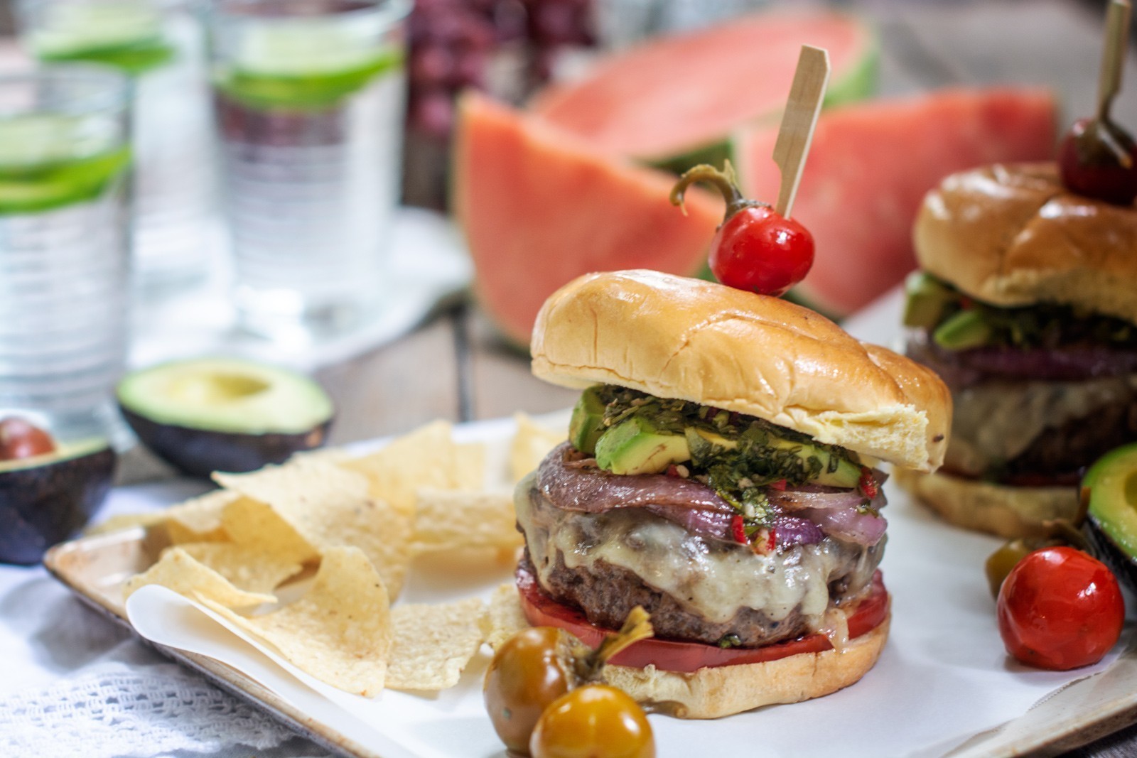 A Chimichurri Burger Recipe features a cheeseburger topped with onions, avocado, and herbs, served with tortilla chips. Cherry tomatoes and avocado halves add freshness, while watermelon and glasses of water complete the scene.