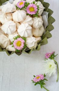 A green bowl filled with easy meringue cookies, decorated with pink and white daisy-like flowers on top and beside the bowl, set on a light wooden surface.