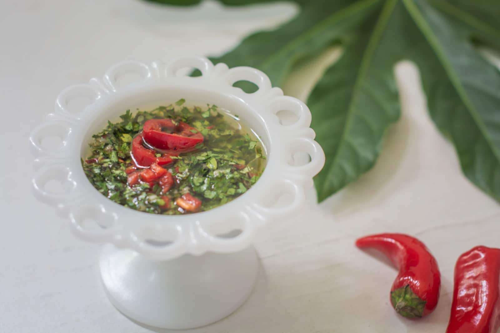 A white ornate cup holds a green herb sauce garnished with sliced red chili peppers, placed on a white surface with two whole red chilies and a large green leaf nearby.