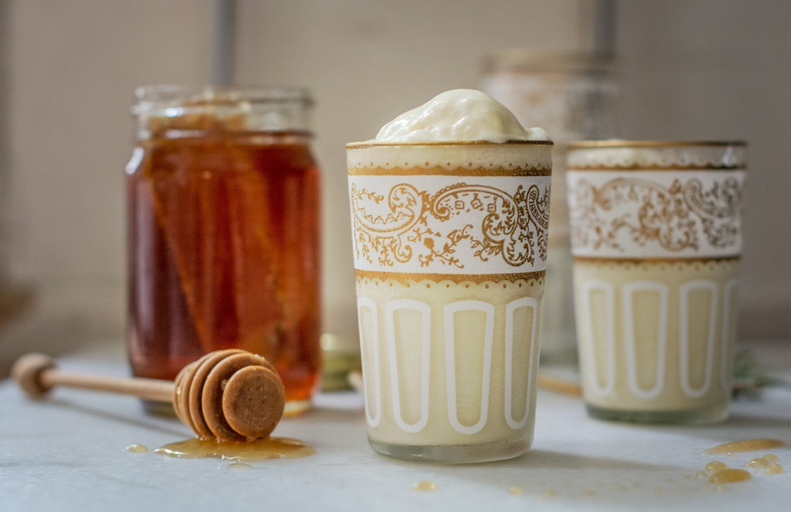 Two ornate glasses filled with a creamy Honey Ice Cream drink, one topped with foam, are placed beside a jar of honey and a wooden honey dipper, with golden honey drips visible on the surface.