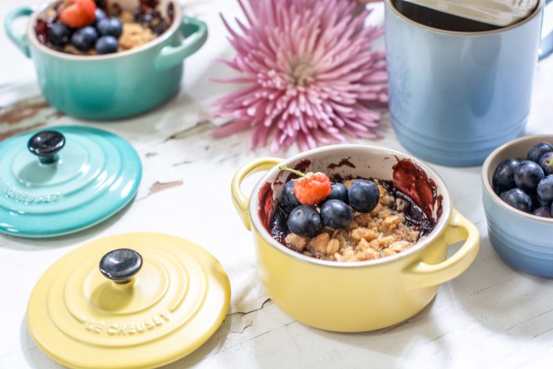 Raspberry and blueberry cobbler in yellow Le Creuset dish on white table and pink flowers