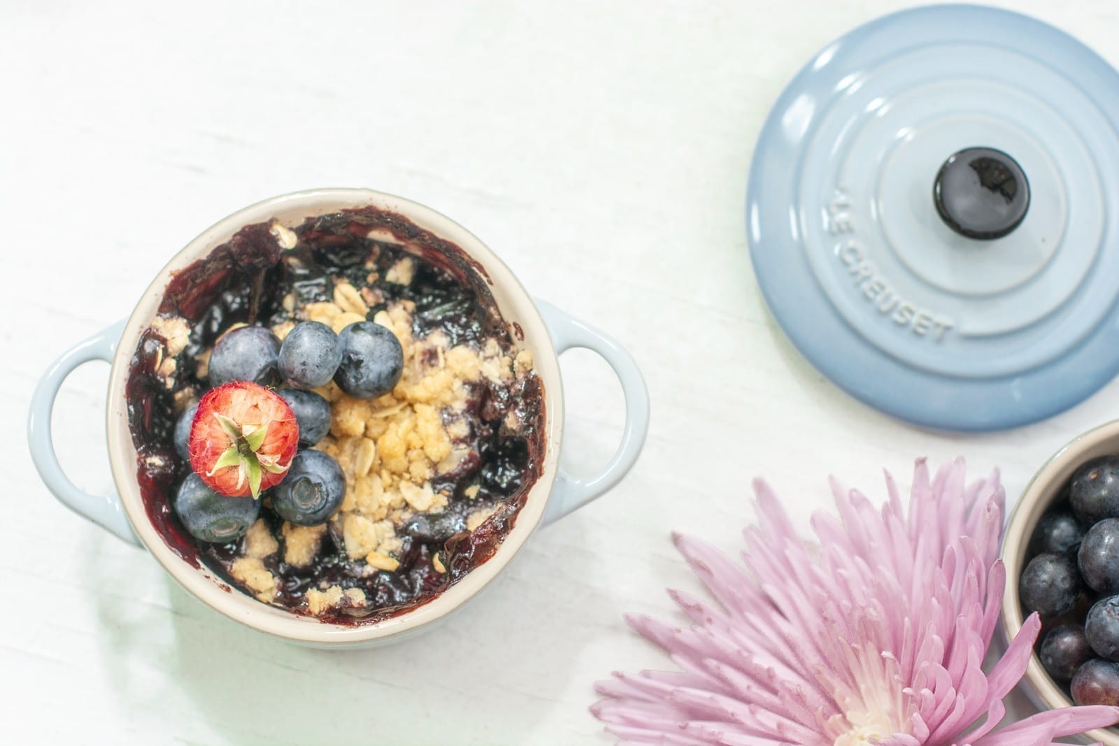 A small blue ceramic pot filled with berry crisp, topped with fresh blueberries and a halved strawberry, sits next to its lid and a pink flower on a light surface.