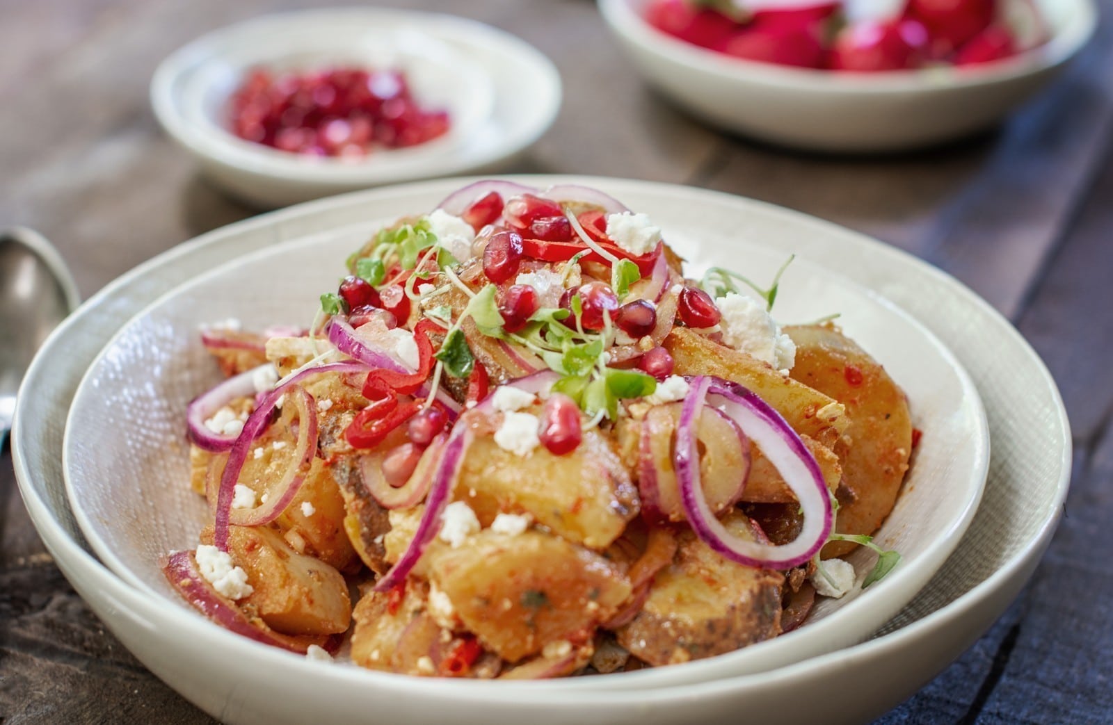 A bowl of potato salad recipe topped with sliced red onions, pomegranate seeds, microgreens, and crumbled cheese, with blurred bowls of extra salad and pomegranate seeds in the background.
