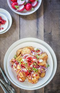 A vibrant potato salad recipe featuring roasted potatoes topped with sliced red onions, pomegranate seeds, crumbled cheese, and microgreens on a wooden table, surrounded by bowls of fresh radishes and extra pomegranate seeds.