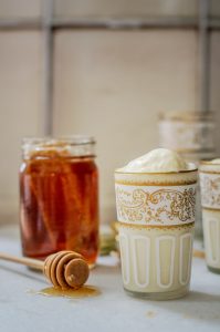 A glass of creamy yogurt with a gold decorative pattern sits beside a jar of honey and a honey dipper on a white surface. Reminiscent of Honey Ice Cream, honey is drizzled around the dipper, adding a touch of sweetness to the scene.