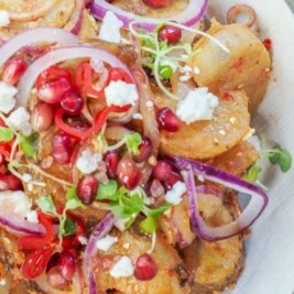 A bowl of Moroccan potato salad recipe topped with red onions, pomegranate seeds, and herbs sits on a wooden table. Nearby are bowls of radishes and extra pomegranate seeds, with a fork and spoon next to the main dish.
