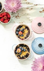 Two small Le Creuset ramekins, one pink and one blue, filled with berry crisp and topped with strawberries and blueberries, sit on a white surface near two large pink flowers and a bowl of fresh raspberries.