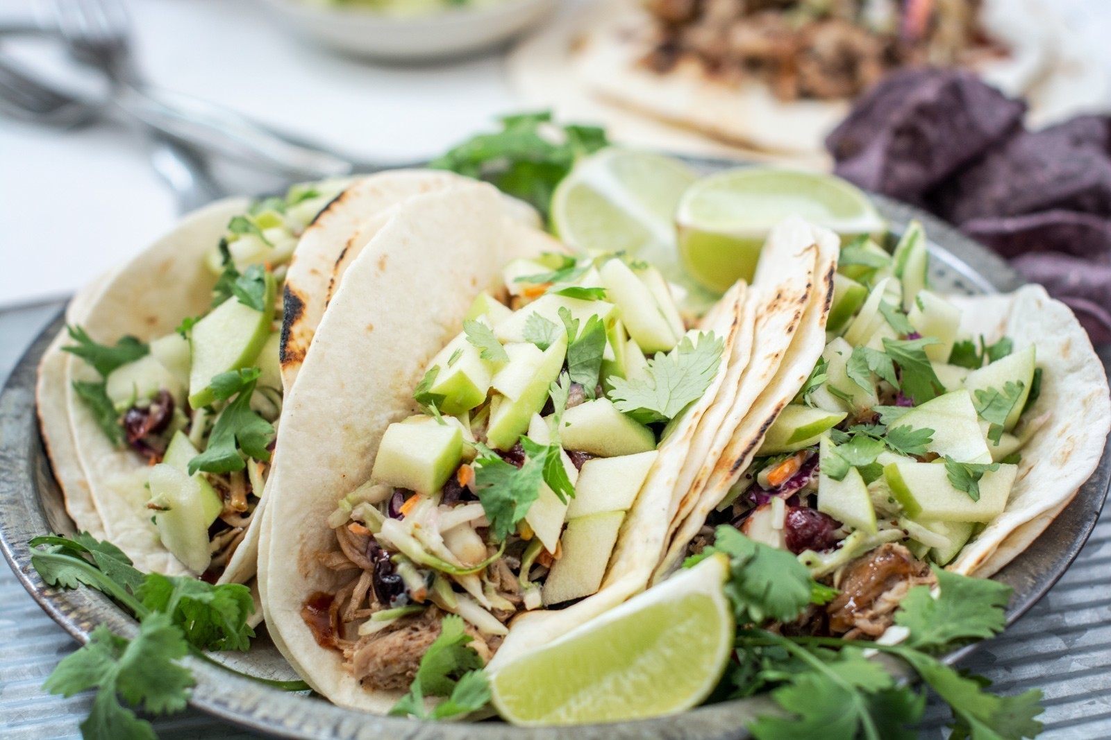 Three soft tacos filled with shredded meat, chopped green apples, cilantro, and other fresh toppings are served on a plate with lime wedges and fresh herbs—a perfect pulled pork taco recipe—with blue corn chips in the background.