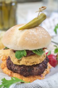 A Southern Burger featuring a beef patty, fried green tomato, bacon, greens, and orange sauce in a bun, topped with pickled okra and secured with a toothpick. Greens and radish garnish are visible around the plate.