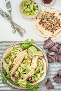 Three soft tacos filled with shredded meat, slaw, diced cucumber, and cilantro are on a plate with lime wedges. Blue corn tortilla chips are on the side—perfect inspiration for your next pulled pork taco recipe.