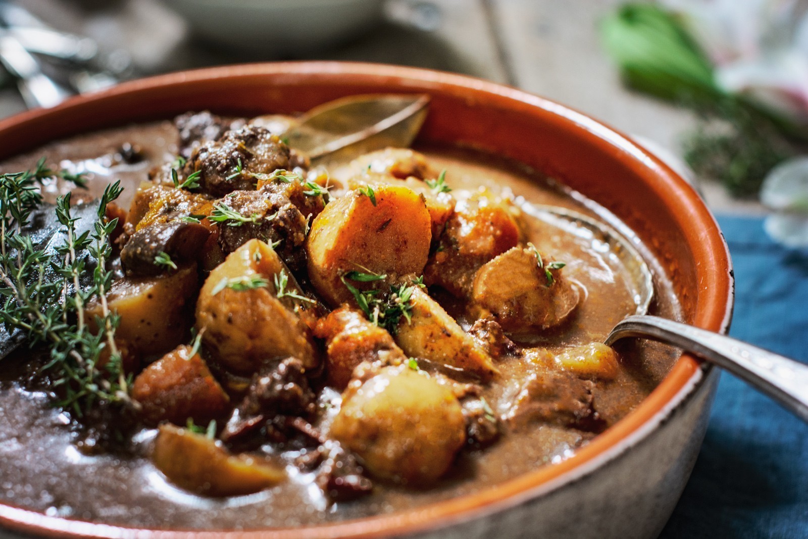A close-up of a hearty beef bourguignon stew with chunks of beef, potatoes, carrots, and herbs in a brown ceramic bowl, garnished with fresh thyme and served with a silver spoon.