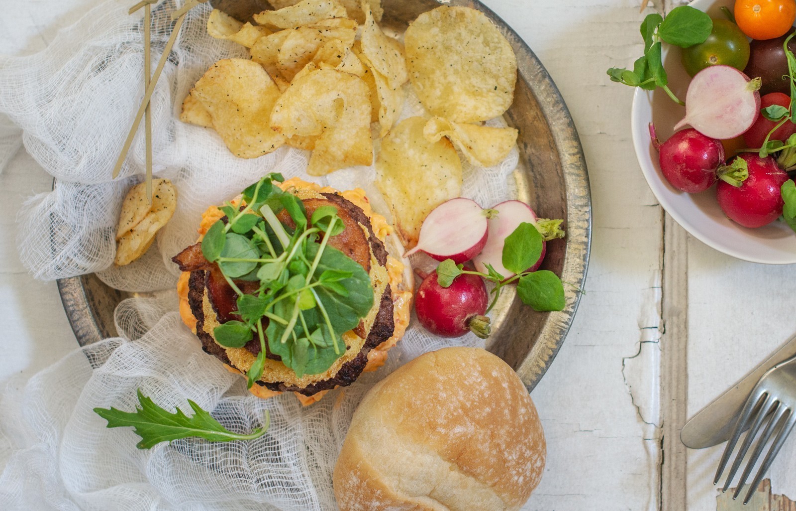 A plate with a Southern Burger veggie patty topped with greens, potato chips, and sliced radishes. Nearby are a bowl of fresh radishes and tomatoes, a sandwich bun, and cutlery on a rustic white surface.