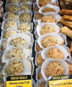 Crab cakes displayed in rows on a deli counter, each sitting in a white paper liner. Signs in front read “Ultimate Lump Crab Cakes” and “Maryland Crab Cakes,” alongside popular crab fries, with price tags below.
