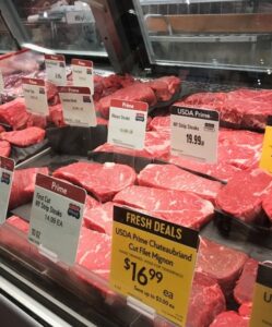 A grocery store meat counter displays various cuts of USDA Prime beef, including strip steaks, filet mignon, and stuffed flank steak, with price tags and Fresh Deals signs visible on the glass case.