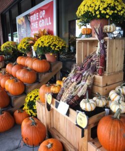 A grocery store display features pumpkins, ornamental gourds, yellow mums, and Indian corn arranged on wooden crates and steps. A Fire Up the Grill sign is visible in the background next to a tempting basket of crab fries.