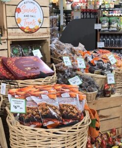 A grocery store display features baskets of autumn-themed treats, including pumpkin pecan granola, crab fries, and chocolate, decorated with a sign reading Autumn Sweets and Fall Treats. Price tags are visible on various items.