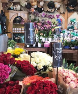 A flower shop displays colorful bouquets of roses with price signs, surrounded by various flowers in pots and baskets—and a small sign for crab fries—arranged on shelves and walls in the background.