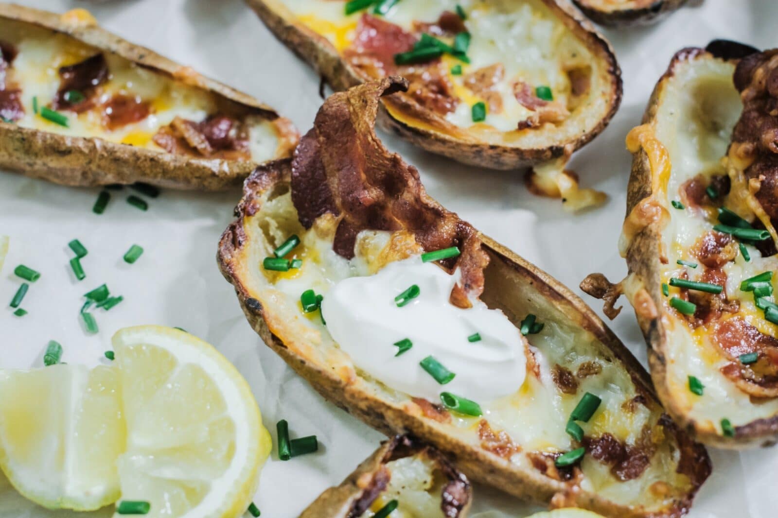 Close-up of loaded potato skins topped with melted cheese, bacon, sour cream, and chopped chives, served with lemon wedges on the side.