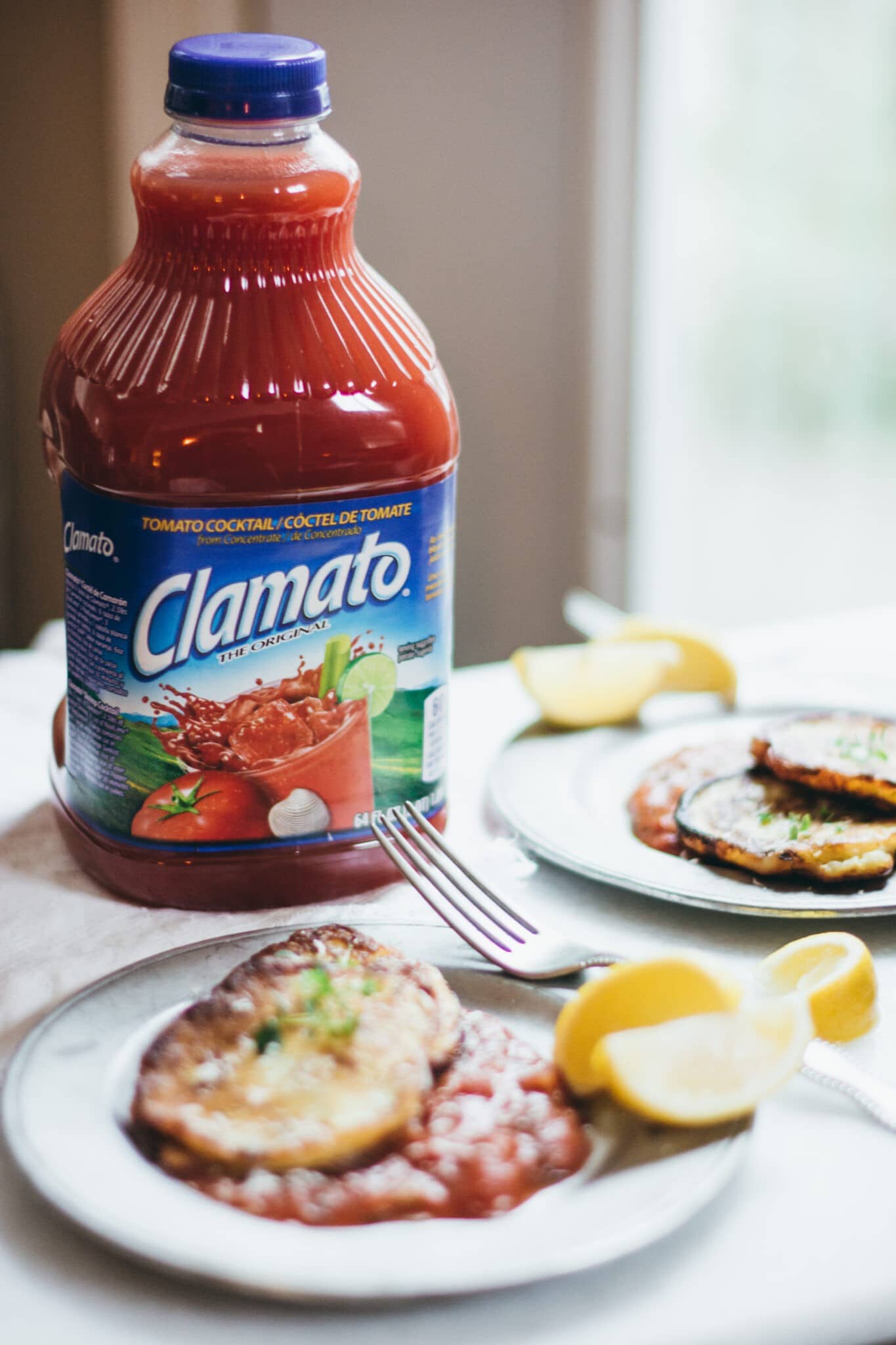 A bottle of Clamato tomato cocktail sits on a table next to two plates of ricotta cakes topped with sauce, garnished with lemon wedges and herbs, with a fork on one plate.