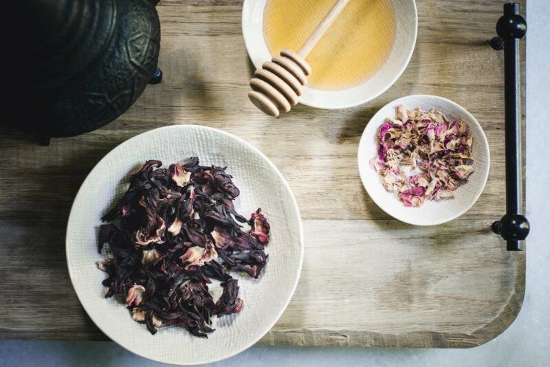 A wooden tray holds a bowl of dried hibiscus petals for Hibiscus Flower Tea, a small bowl of dried rose petals, and a dish of honey with a honey dipper. The items are arranged neatly for tea preparation.
