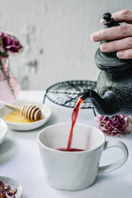 A hand pours vibrant Hibiscus Flower Tea from a black teapot into a white cup on a white table, with a honey dipper, pink flowers, and a decorative plate in the background.