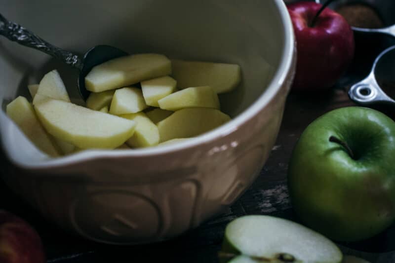 A ceramic bowl filled with sliced apples and a spoon sits on a wooden table, surrounded by whole apples and measuring cups—perfect for preparing a homemade apple cobbler.