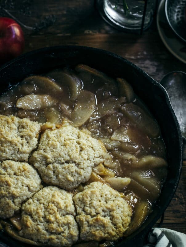 A close-up of a skillet filled with apple cobbler, displaying golden-brown biscuit topping over warm, caramelized apple slices in syrup. A spoon and glass are partially visible in the background, highlighting this classic Apple Cobbler dessert.
