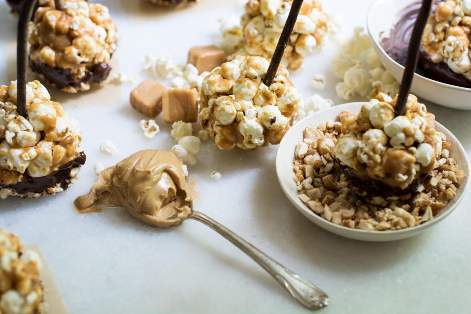 Popcorn balls on sticks are coated with caramel and chocolate, served alongside bowls of chopped nuts and chocolate for dipping. A spoonful of peanut butter and caramel candies are scattered around the popcorn balls on a white surface.
