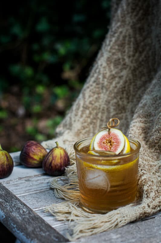 A glass of amber-colored rum cocktail with ice is garnished with a fig slice and lemon twist. Fresh figs rest nearby on a rustic wooden surface, with a textured beige cloth draped in the background.