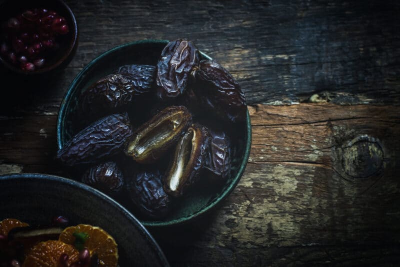 A bowl of dark brown dates, with one date split open to reveal its sticky interior, sits on a rustic wooden surface. Nearby are hints of Orange Salad with pomegranate seeds and orange slices in other dishes.