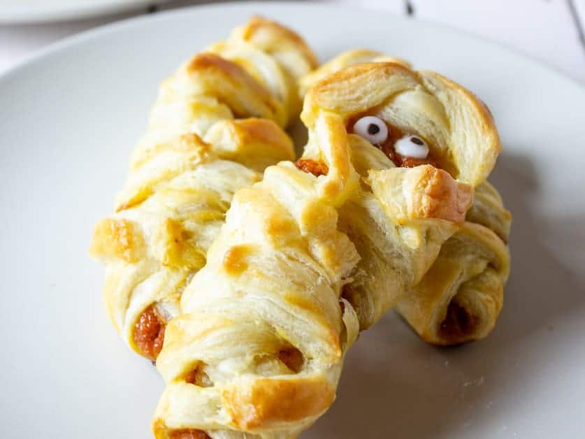 Two puff pastry mummies with candy eyes and braided, golden-brown dough, placed on a white plate. The pastries are designed to look like mummies for a Halloween-themed treat.