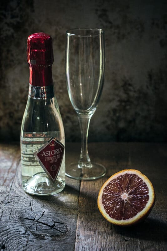 A small bottle of Astoria Prosecco, an empty champagne flute, and a halved blood orange sit on a rustic wooden surface, ready to inspire your next bellini recipe against a textured, dimly lit background.