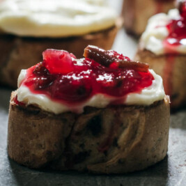 A close-up of a pastry slice topped with creamy white frosting and glossy, chunky cranberry jam, resting on a gray surface. Other similarly frosted pastries are blurred in the background.