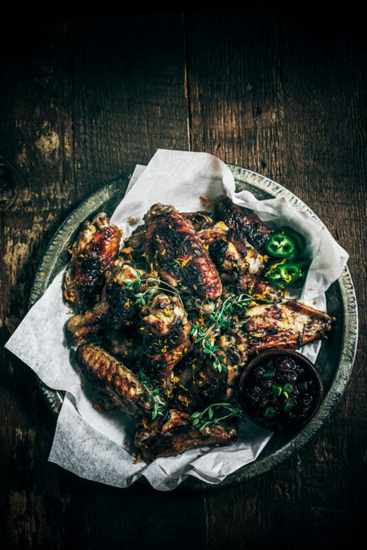 A rustic plate lined with parchment paper, piled high with sticky chicken wings garnished with fresh herbs, served with a small bowl of dark dipping sauce and sliced green jalapeños on the side.