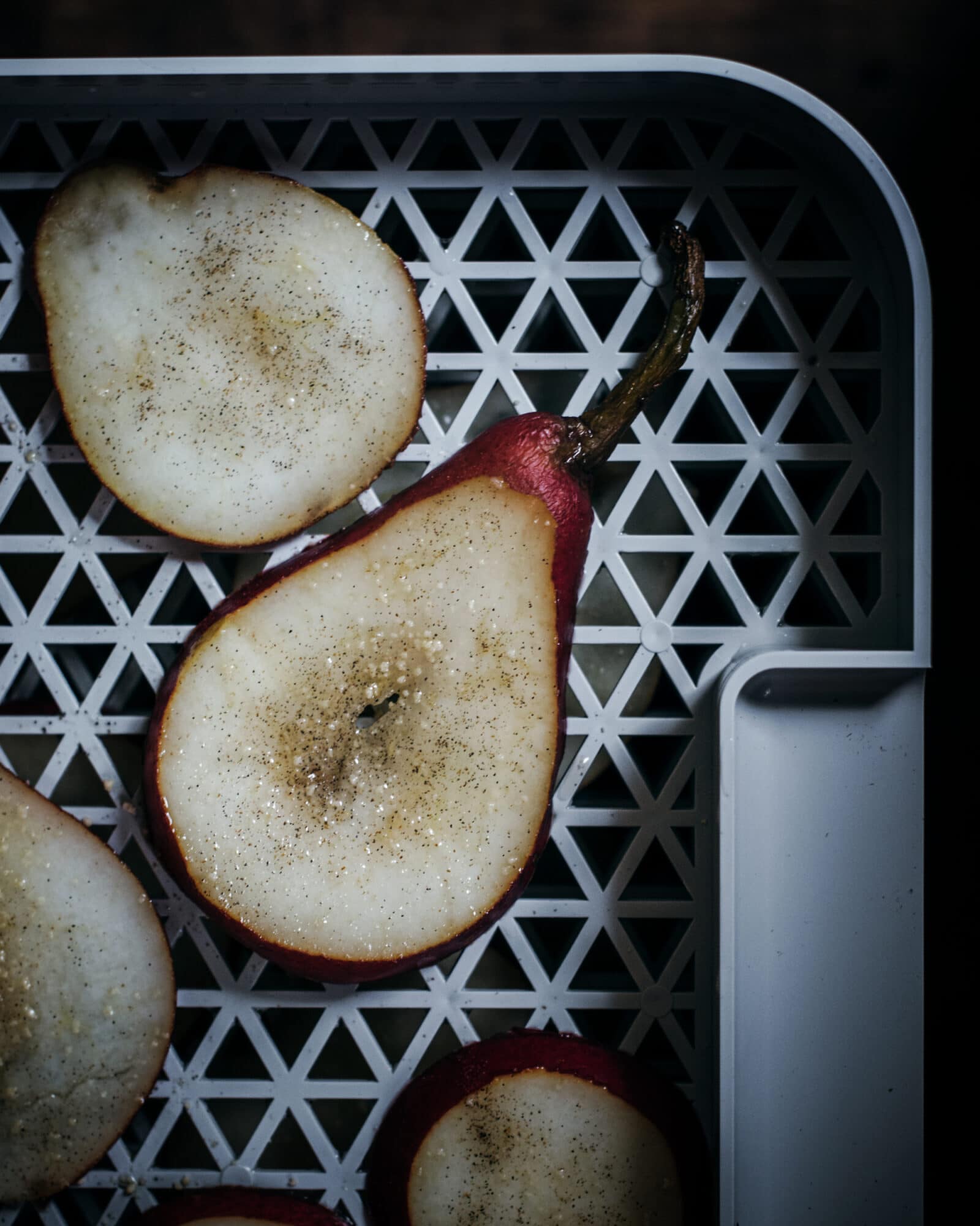 A sliced pear with a red skin sits on a white, patterned tray, sprinkled with sugar and spices. Other pear slices are partially visible around it. The lighting is moody and highlights the pear’s texture.