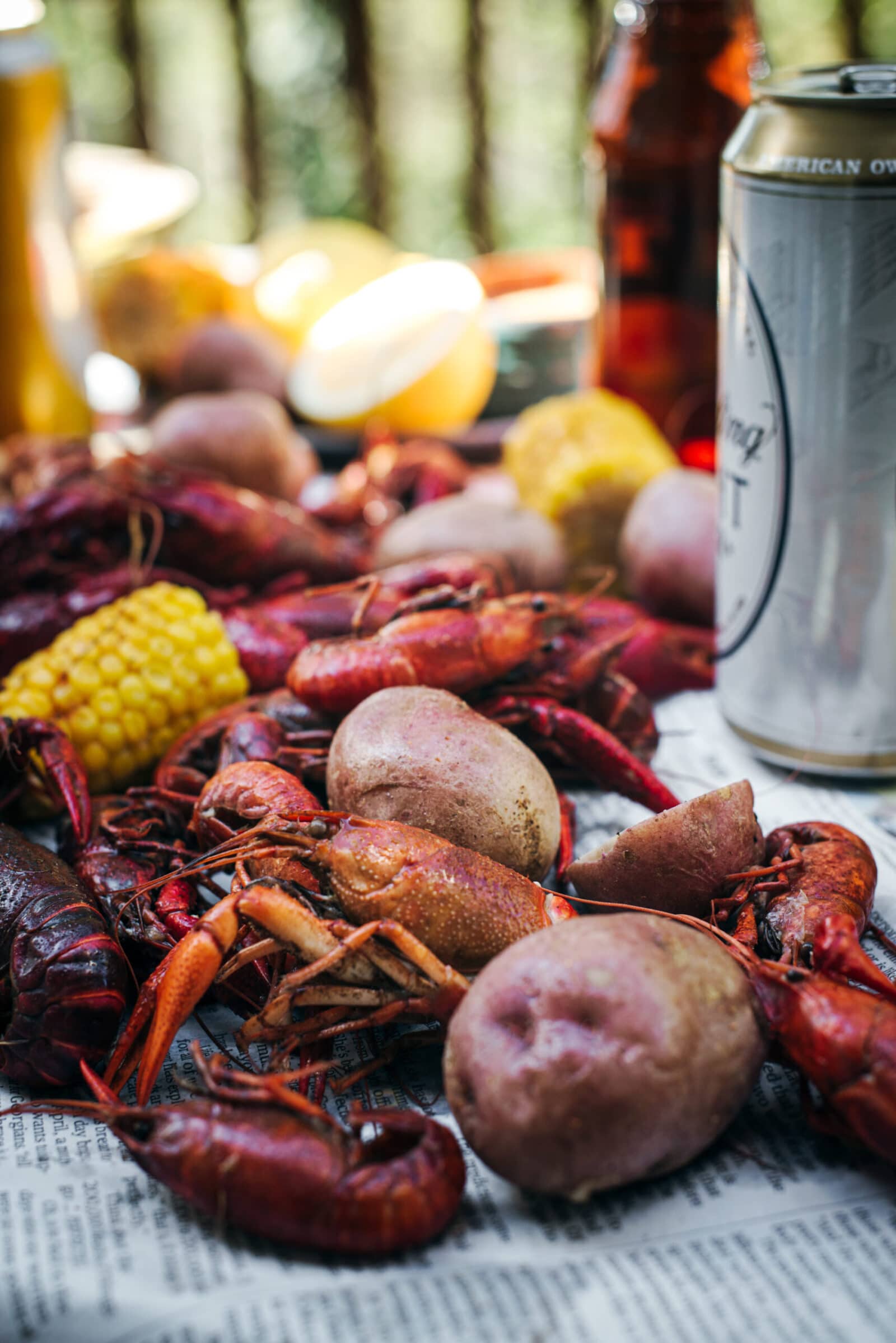 A close-up of a classic crawfish boil spread on newspaper, featuring crawfish, red potatoes, corn on the cob, and beverages in cans and bottles in an inviting outdoor setting.