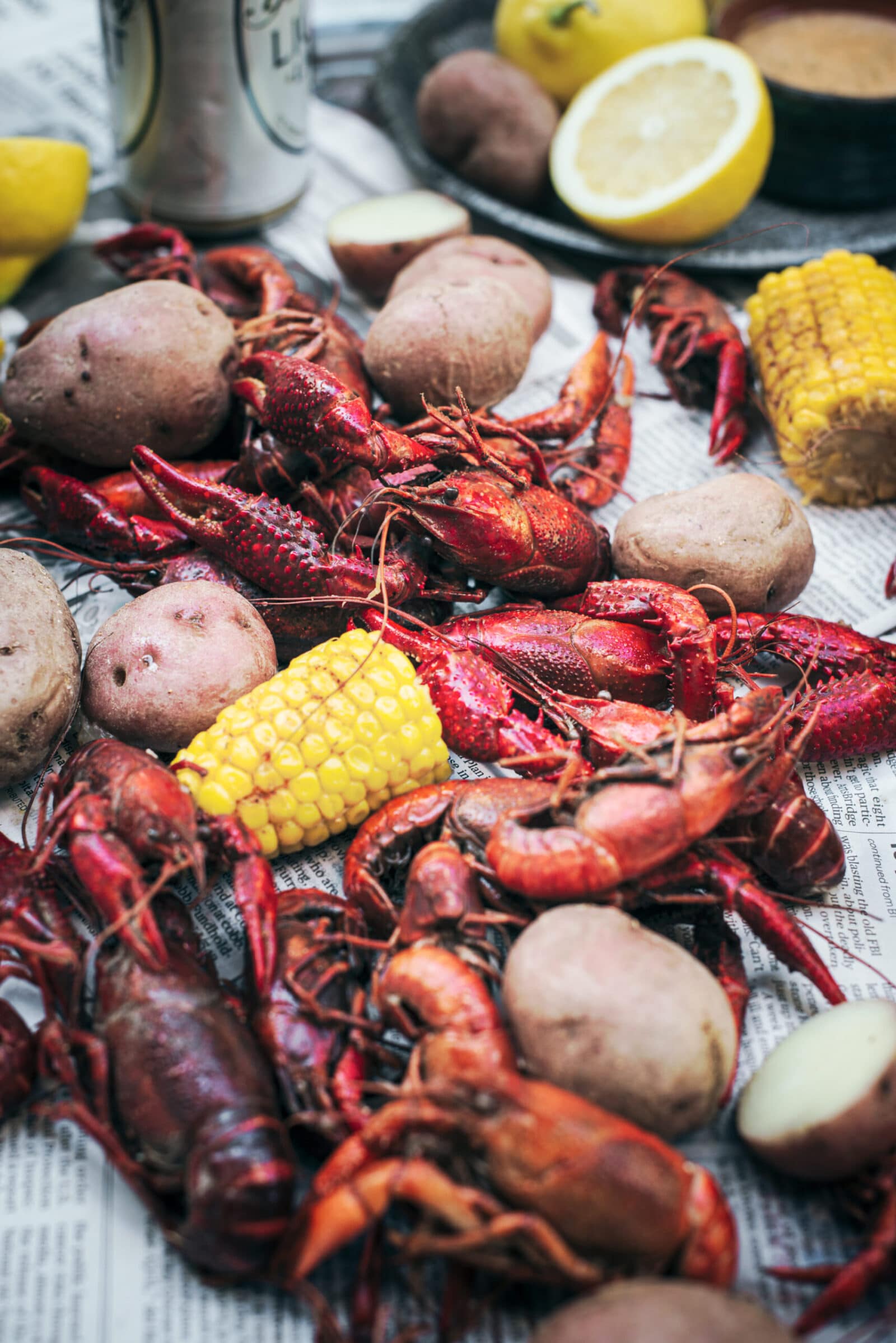 A pile of cooked crawfish, potatoes, and corn on the cob is spread on newspaper, with lemons and drinks in the background, capturing the laid-back vibe of a classic crawfish boil.