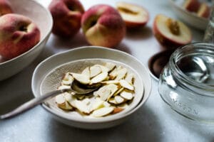 A bowl of sliced almonds with a spoon rests on a table, surrounded by whole and halved peaches, an empty Mason jar, and other bowls—perfect ingredients for creating homemade peach and almond jam.