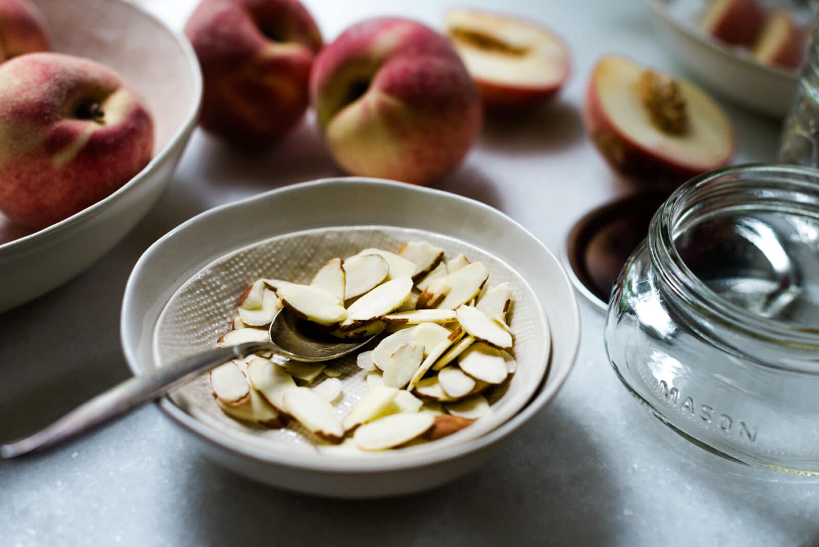 A bowl of sliced almonds with a spoon rests on a table, surrounded by whole and halved peaches, an empty Mason jar, and other bowls—perfect ingredients for creating homemade peach and almond jam.