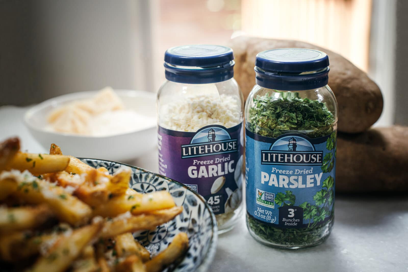 Two jars of Litehouse freeze dried garlic and parsley sit beside potatoes, a bowl of grated cheese, and a plate of truffle fries on a kitchen counter.