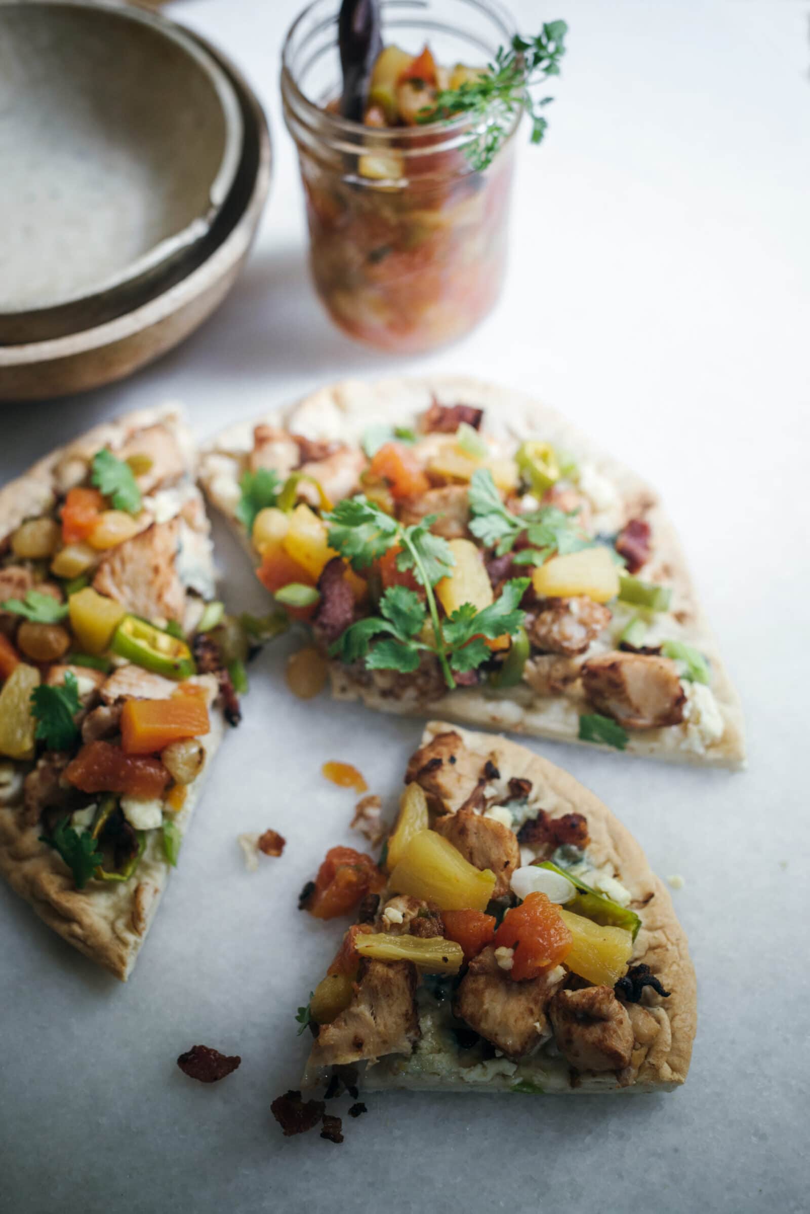 Three slices of pizza topped with chicken, pineapple, vegetables, and herbs are arranged on a white surface, with a jar of salsa recipe and stacked plates in the background.