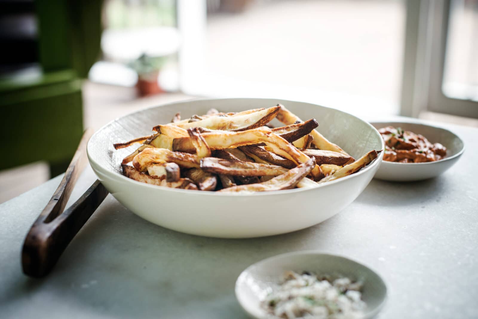 A bowl of golden, crispy Air Fryer Fries sits on a table with wooden tongs resting on the rim. In the background are two small dishes—one with dip and the other with chopped herbs or seasoning, perfect for this Homemade French Fries Recipe.