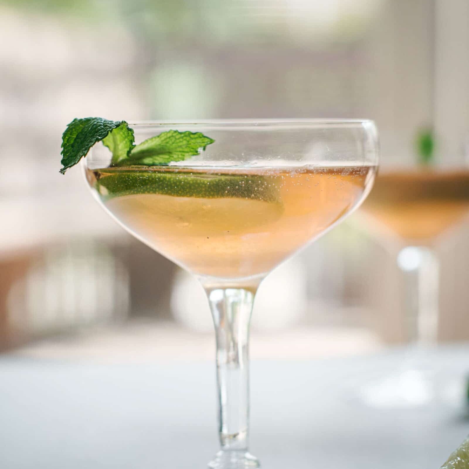 A coupe glass filled with a light pink sparkling old Cuban cocktail, garnished with a lime wedge and fresh mint sprig, sits on a light surface with another similar drink blurred in the background.