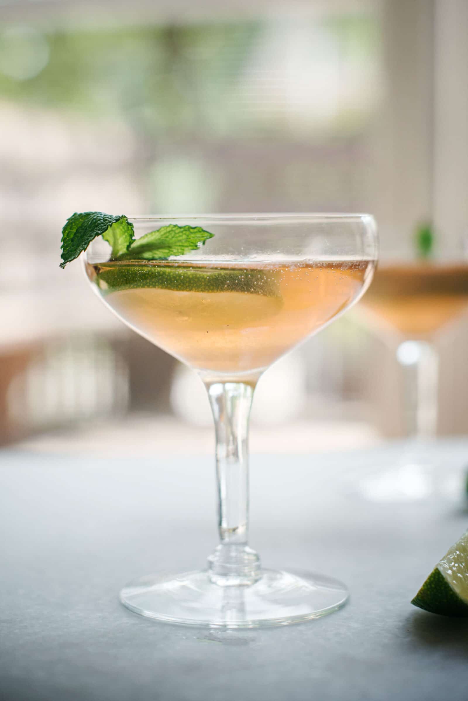A coupe glass filled with a light pink old Cuban cocktail, garnished with a slice of lime and a sprig of mint, sits on a gray surface with a blurred background.