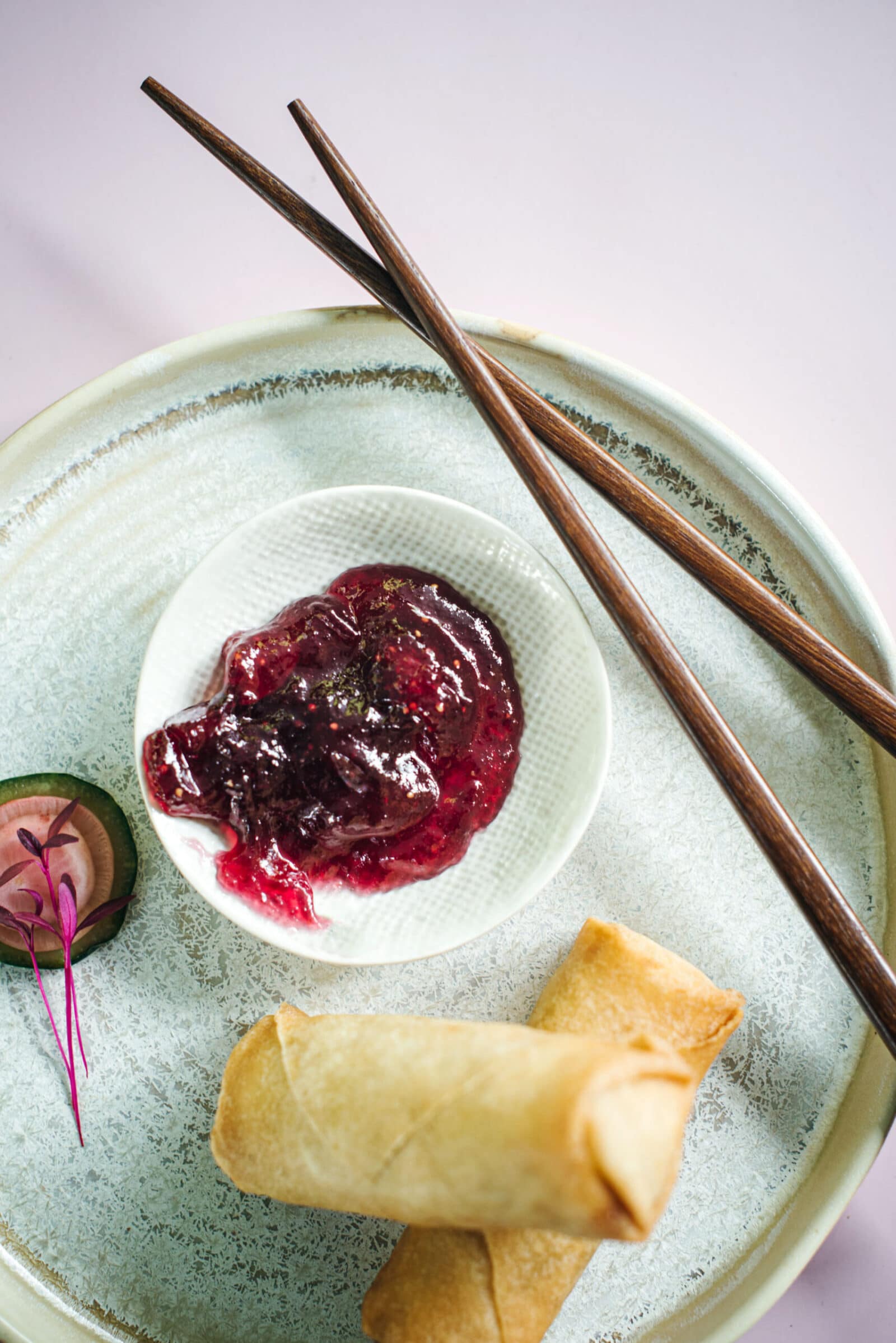 A plate with two spring rolls, a small bowl of red dipping sauce reminiscent of a fig jam recipe, cucumber garnish, and a pair of wooden chopsticks on a light background.